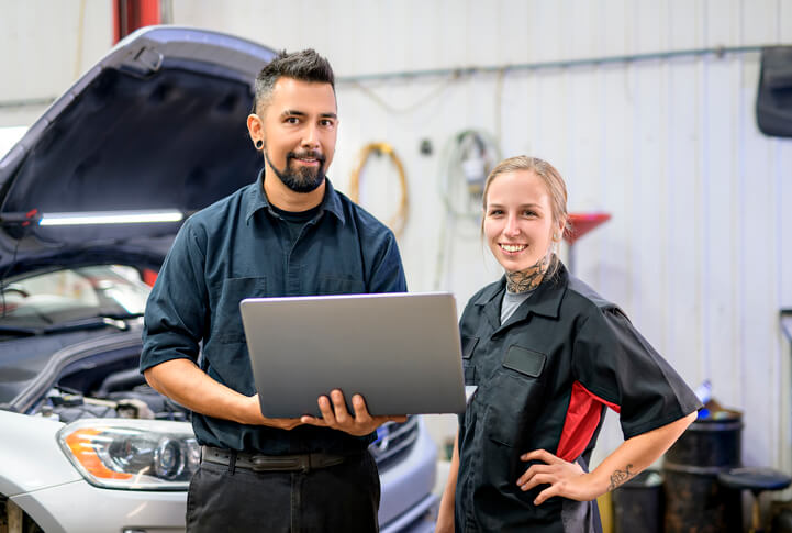 An auto mechanic training grad stands with a mechanic in a shop setting 